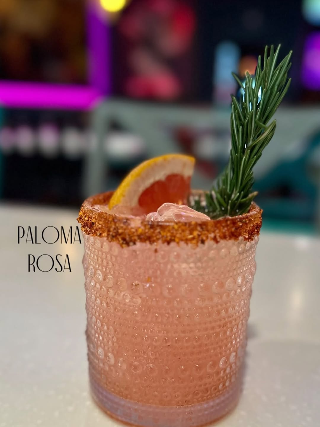 Paloma Rosa — pink cocktail with tajín rim under pink neon glow